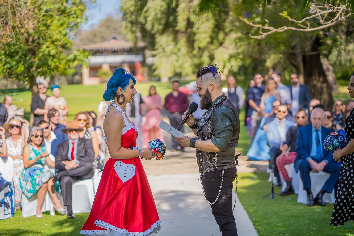 A couple with vibrant blue and red outfits perform a dance at an outdoor wedding ceremony as guests watch from the lawn.