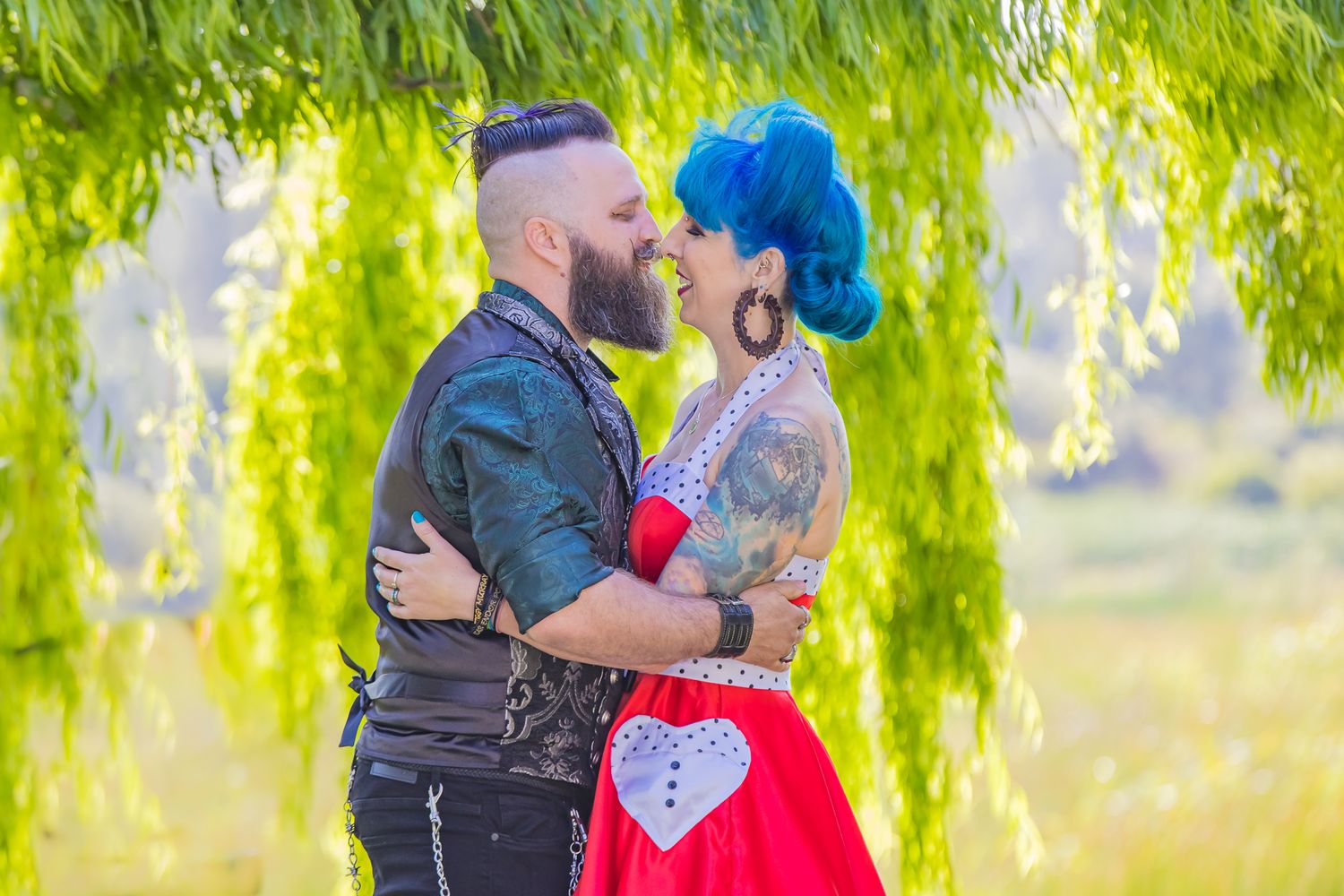 A couple embraces under a weeping willow tree, with the partner wearing a red vintage-style dress and blue hair.