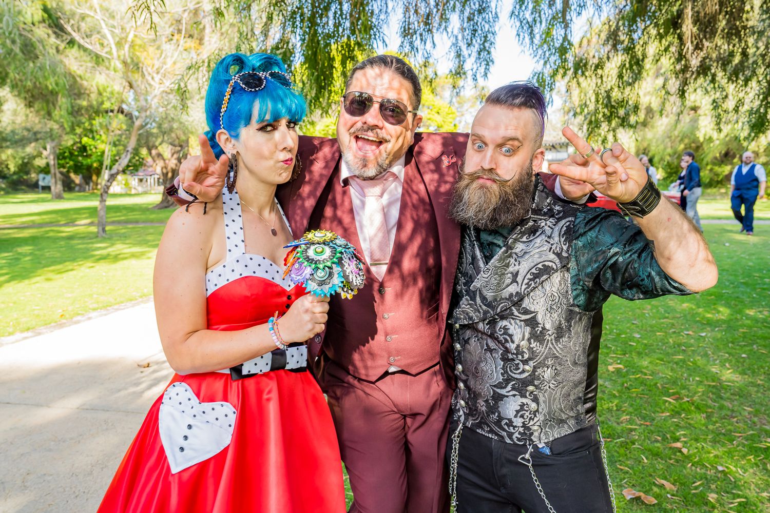 Three friends in colorful outfits pose playfully at an outdoor party in a sunny park setting.