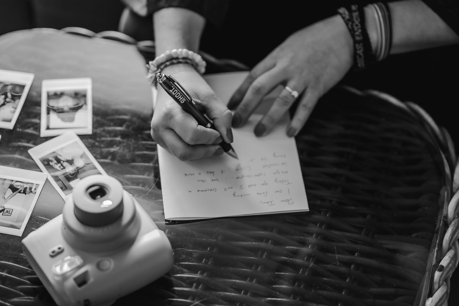 Black and white image of hands writing on paper next to a Fujifilm Instax instant camera and photos on table.