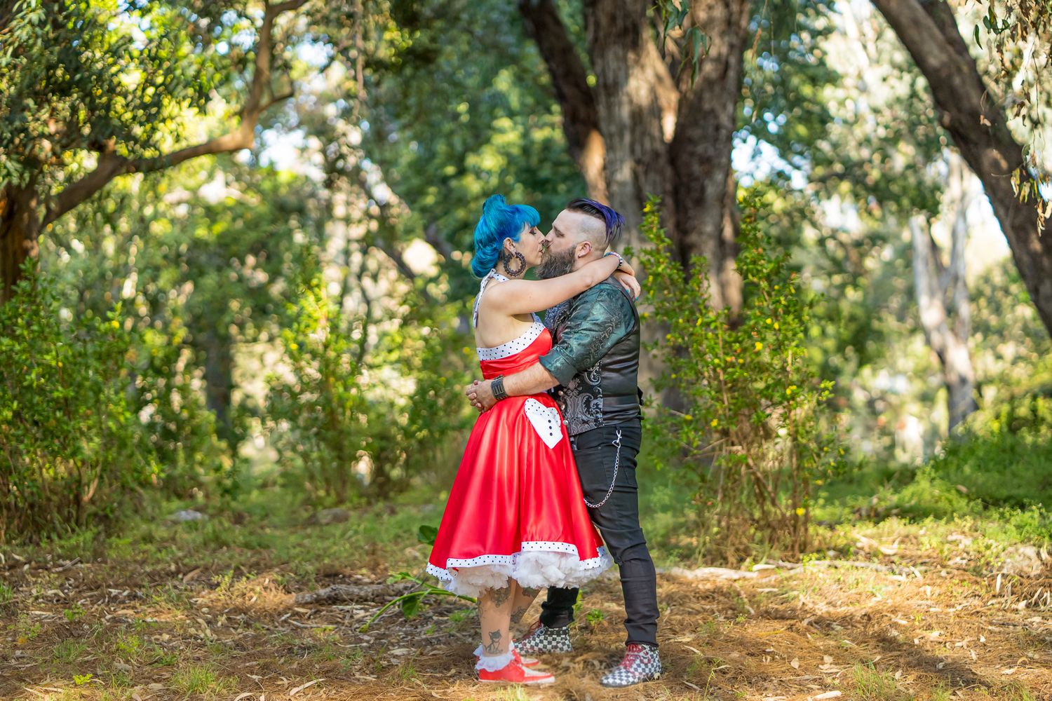 A couple embraces in a sunlit forest clearing, with the woman wearing a vibrant red dress.