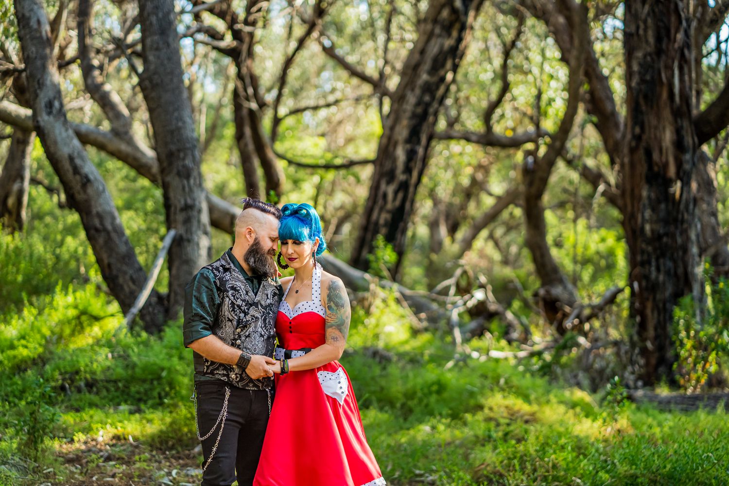 A couple stands together in a forest setting with her wearing a vibrant red dress and him in dark casual attire.
