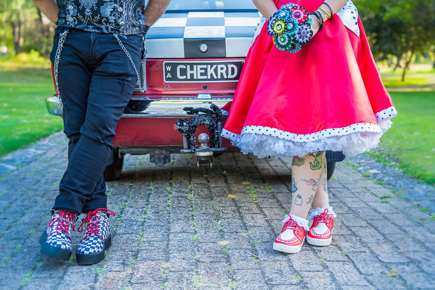 Two pairs of feet wearing matching rockabilly style shoes stand near a vintage car with patterned trim details.