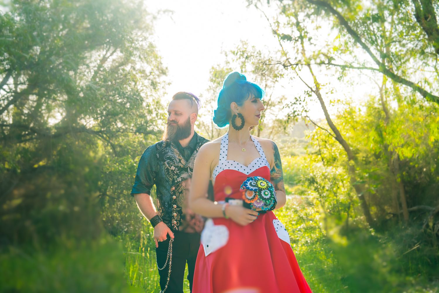 A couple in rockabilly style poses in a sun-drenched garden, with a red dress and guitar creating a vintage aesthetic.