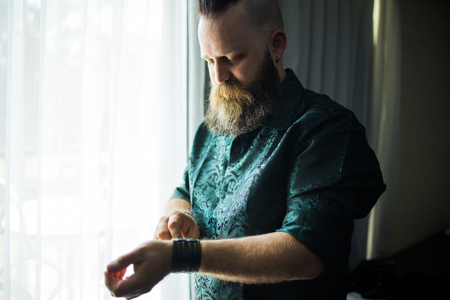 Someone with a large beard wearing a teal patterned shirt checks their smartwatch in natural window lighting.