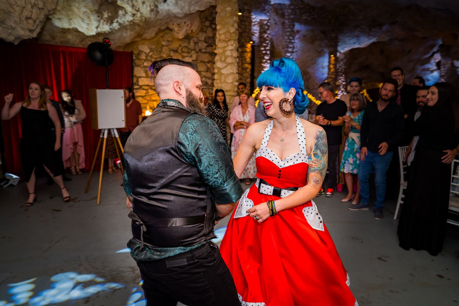Two people dance together at a party, with one wearing a bright red dress and blue hair while the other wears black leather.