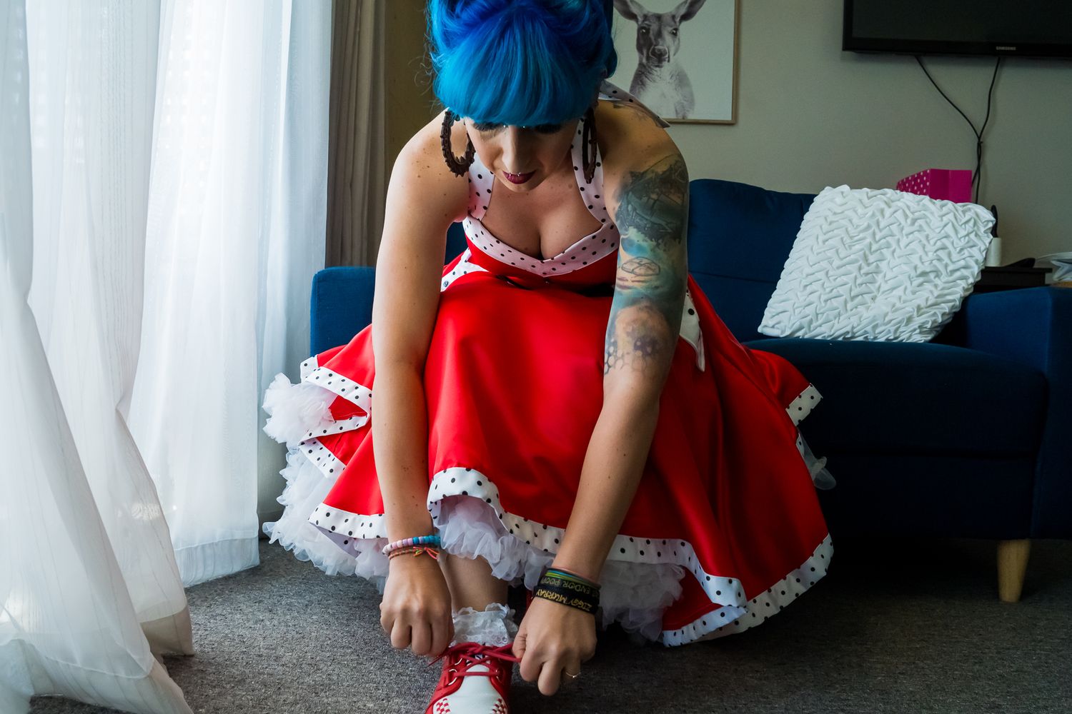 A person in a blue cap adjusts red and white ruffled dress while wearing sneakers in a living room setting.