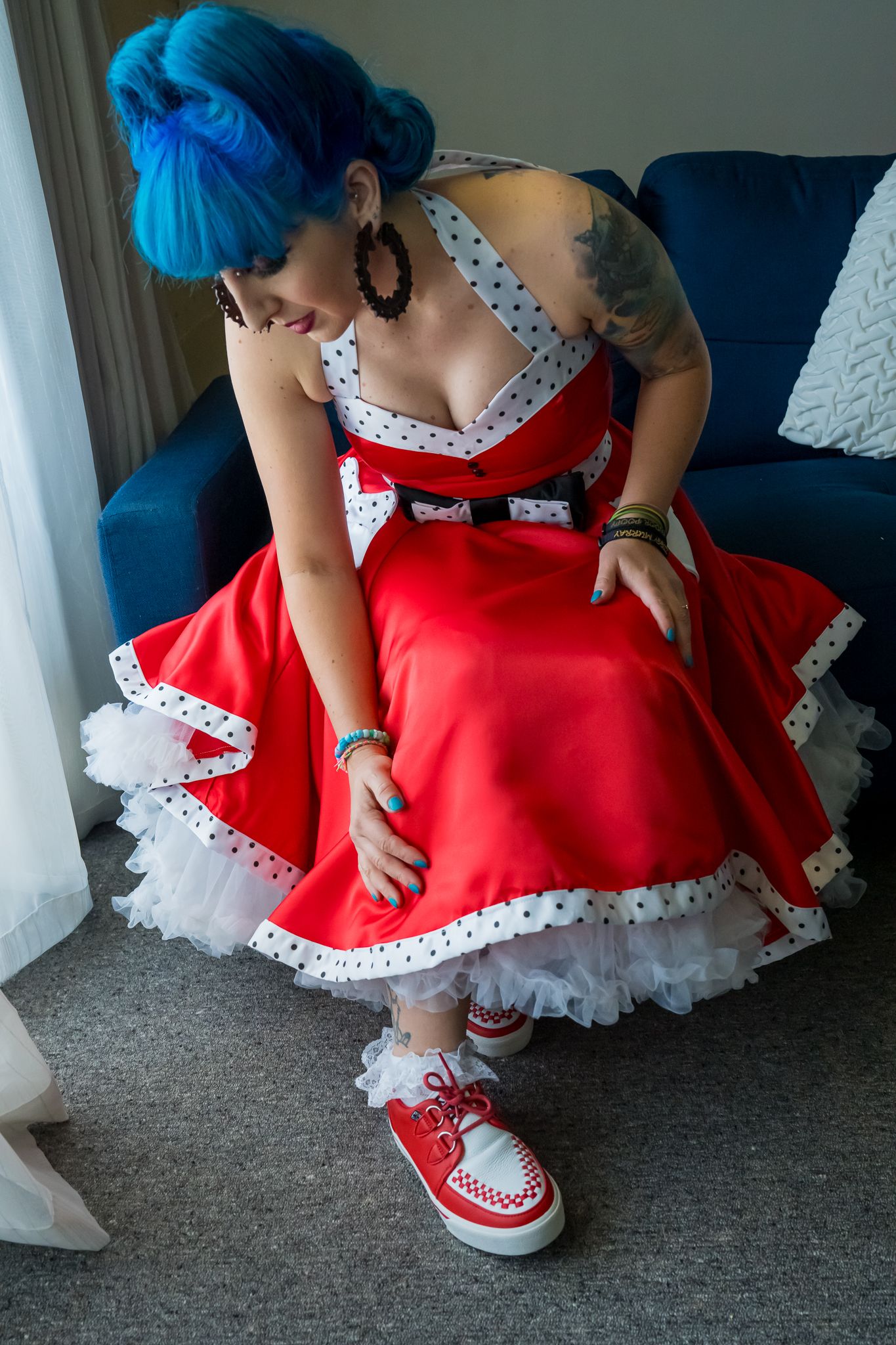 A person in a red and white polka dot rockabilly dress with tulle petticoat and matching red converse sneakers sits on a couch.