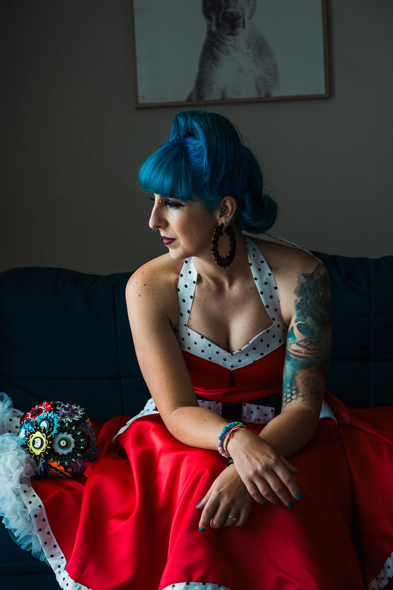 A woman in a red dress with blue hair sits on a sofa with a cat picture visible on the wall behind her.
