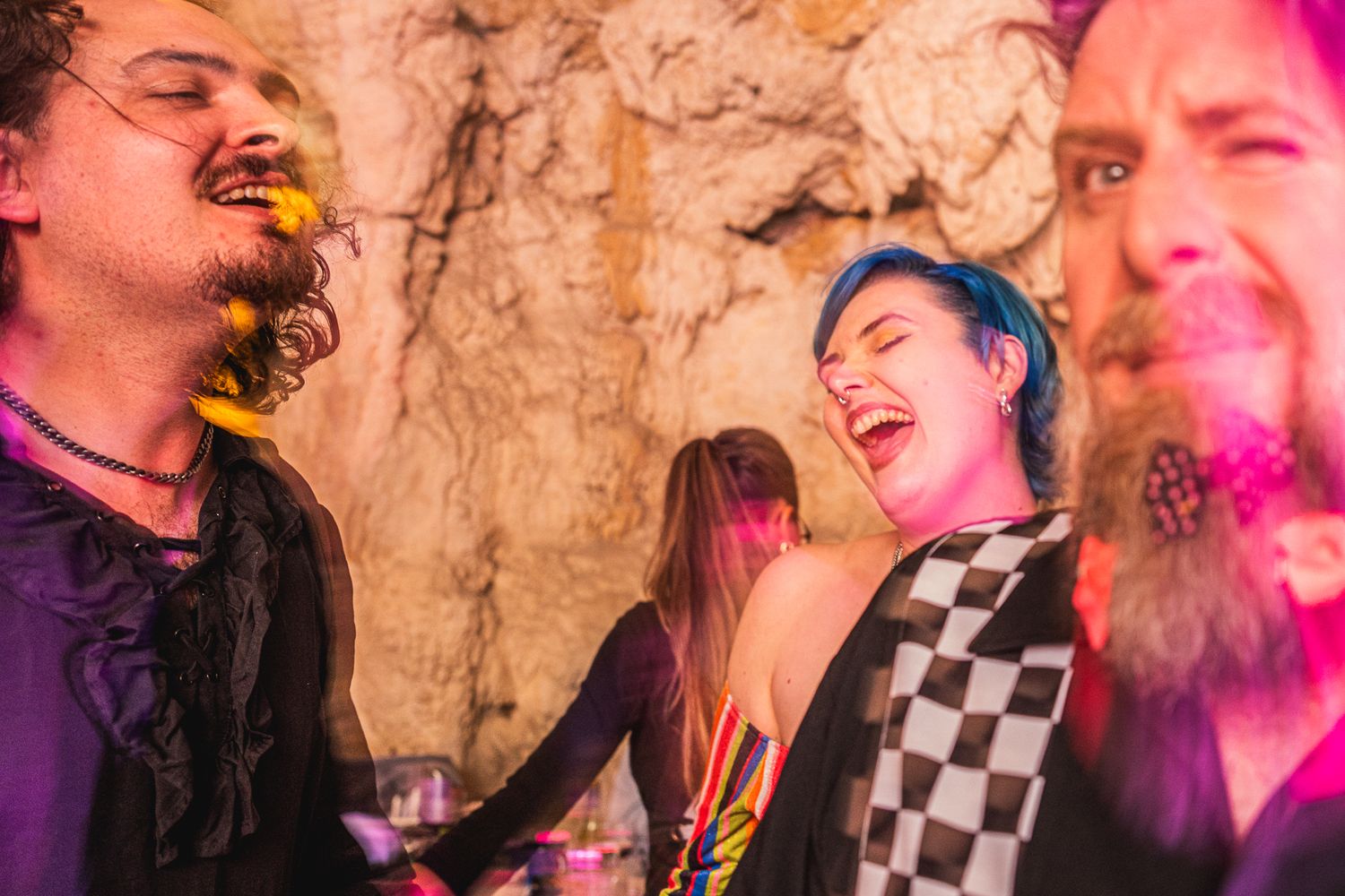 Group of friends laughing together at a party with colorful pink lighting and checkered patterns visible.