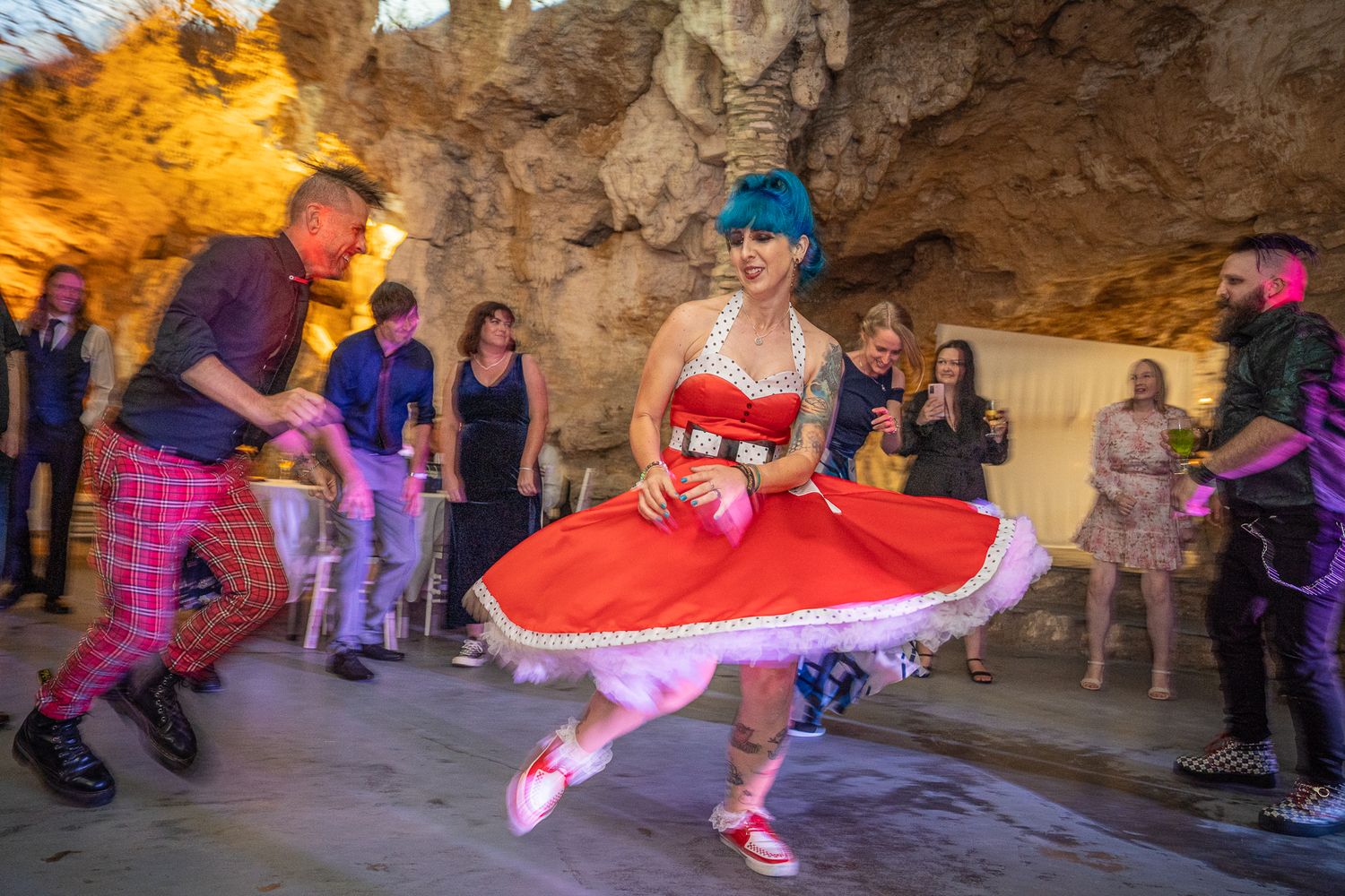 Dancers perform in colorful outfits inside a cave-like setting while spectators watch from the sides.