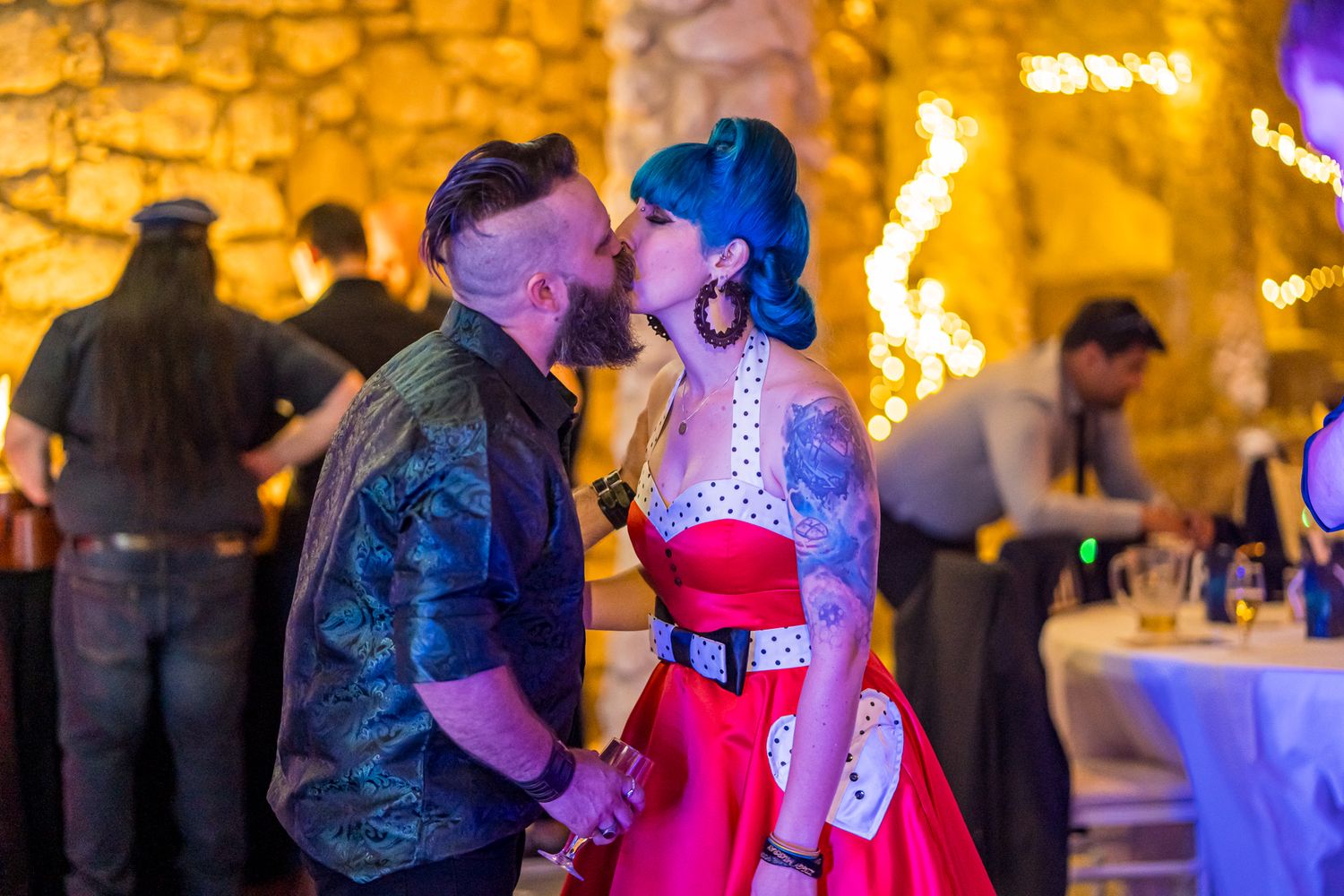 A couple shares a romantic kiss at a party with glowing stone walls and twinkling lights in the background.
