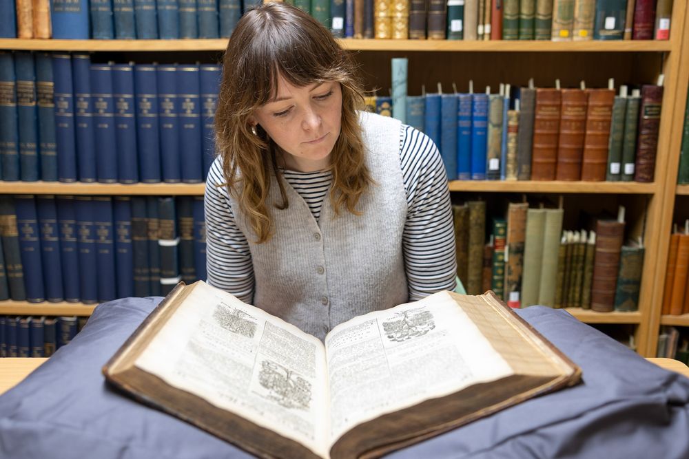 A person in a striped sweater reads an old volume at a library desk with bookshelves visible in the background.