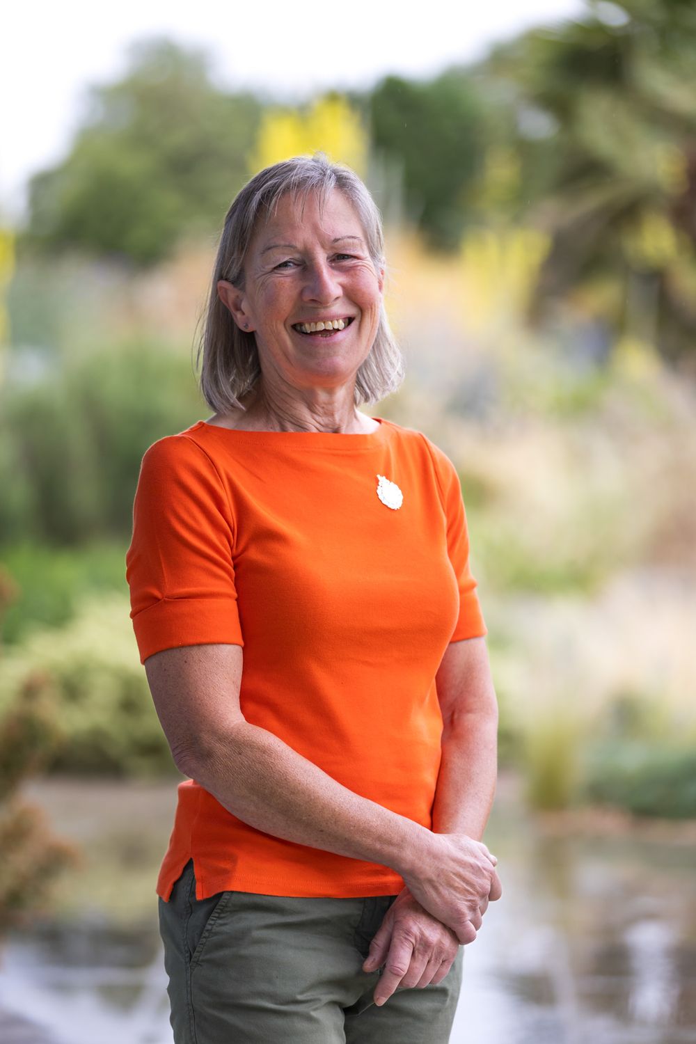 Someone wearing a bright orange t-shirt and olive pants stands outdoors smiling in a natural setting.