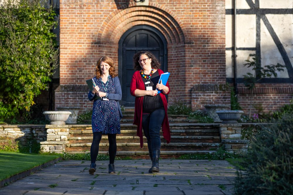 Two colleagues having animated discussion in front of arched brick doorway on collegiate campus.