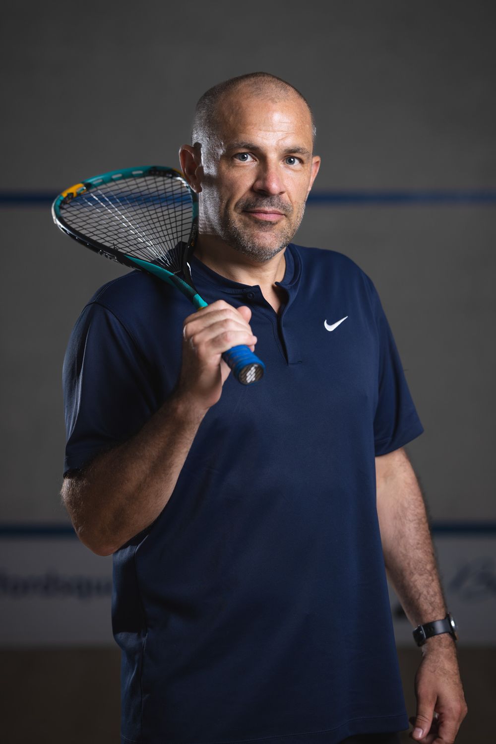 Athletic instructor in navy polo holds tennis racket over shoulder.