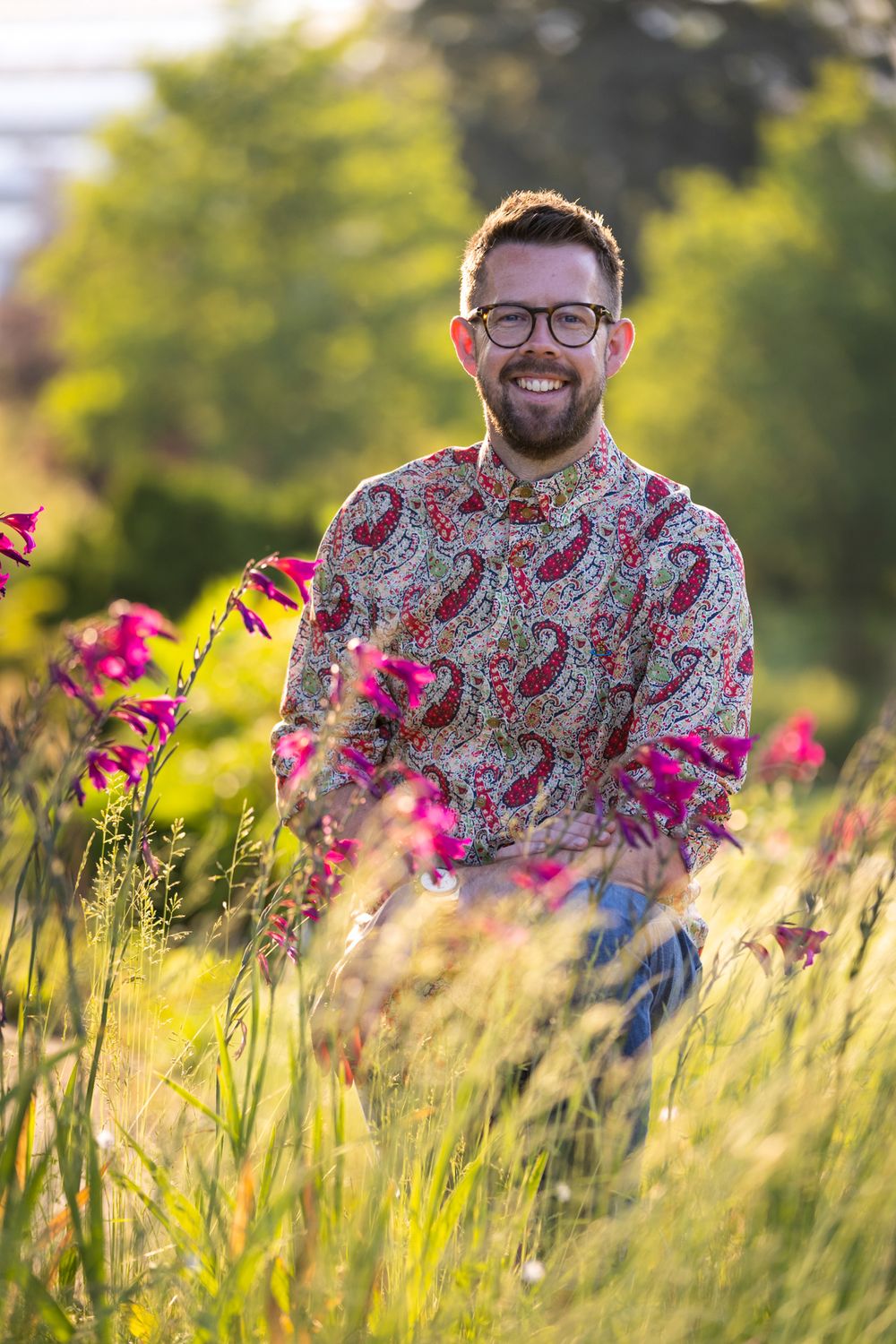 Someone wearing glasses and a paisley print shirt smiles while standing in a field of tall grass and pink wildflowers at sunset.