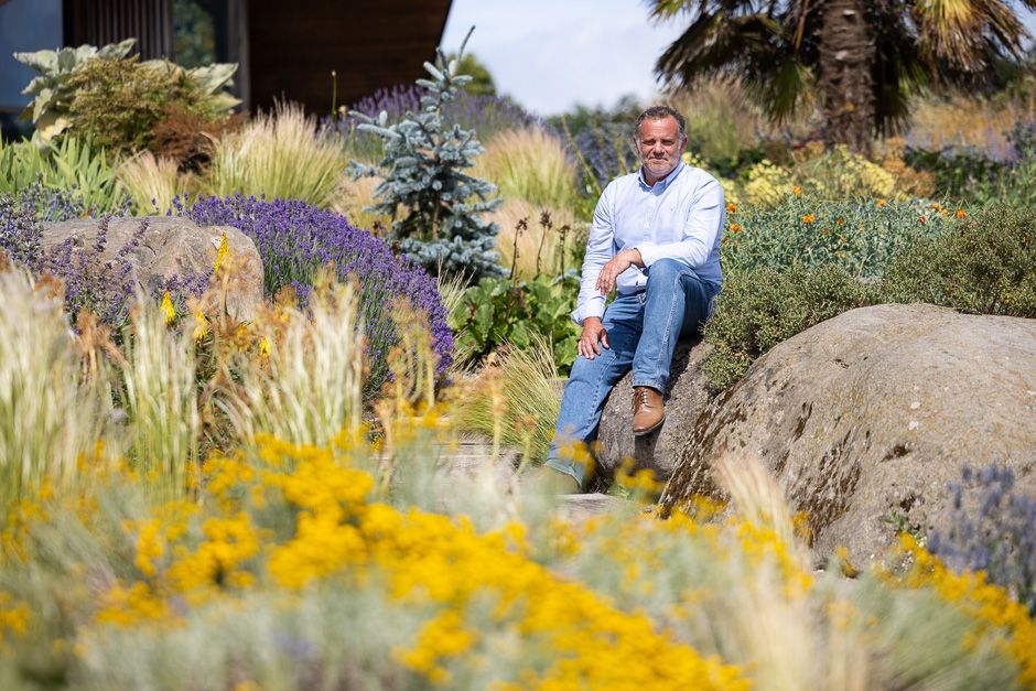 A person sits among colorful perennial flowers and ornamental grasses in a drought-resistant garden landscape.