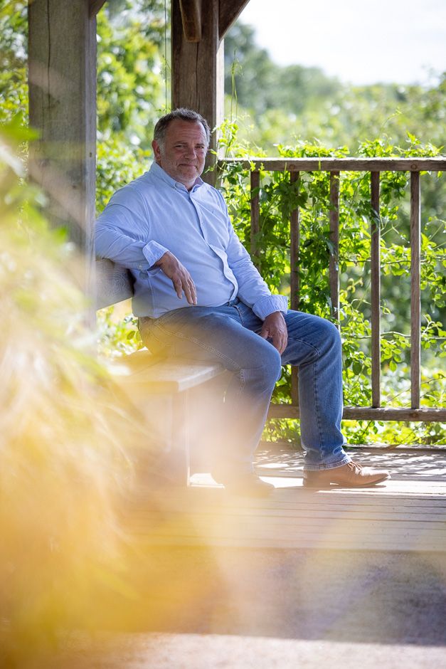 Person sitting alone on a wooden deck with sunlit greenery visible through railing in background.
