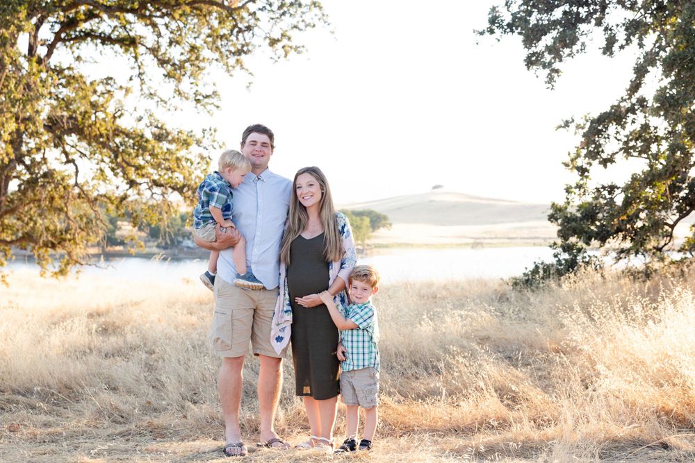 A family of four poses together in a sunny field with golden grass and trees during an outdoor photo session.