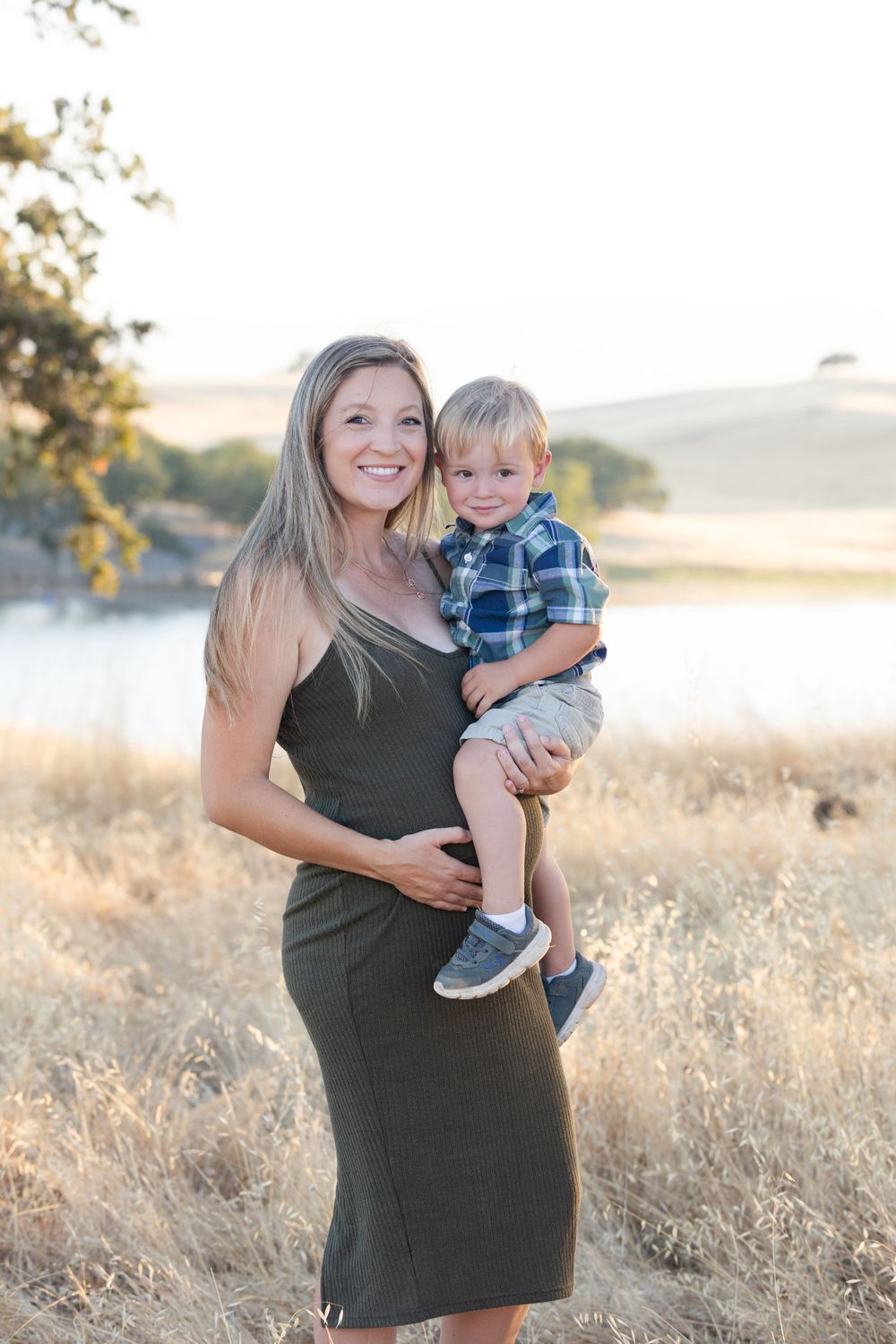 A mother and toddler pose together in an olive dress against a serene golden hillside landscape at sunset.