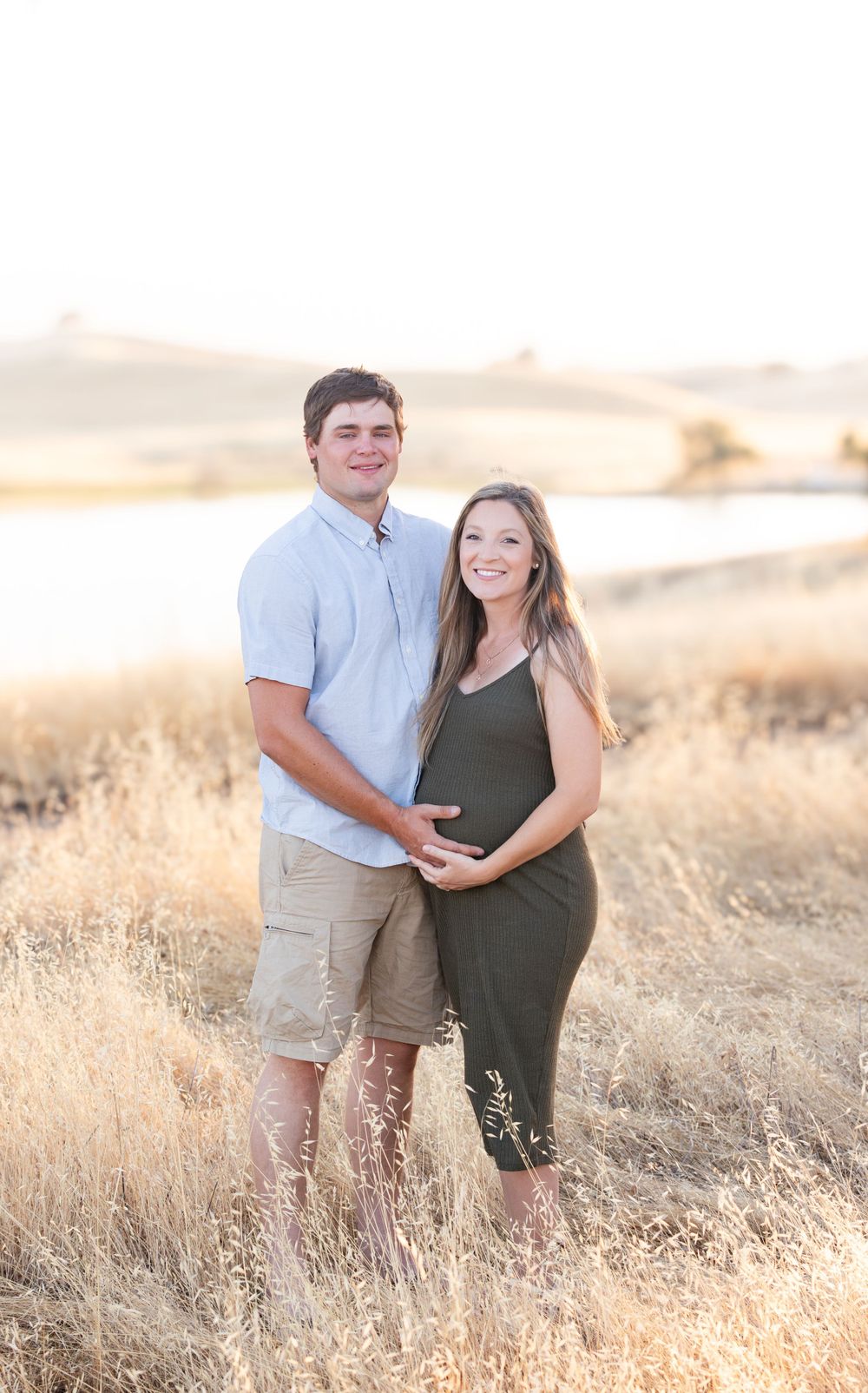 A couple stands together in a golden field during sunset as they pose for a maternity photo.