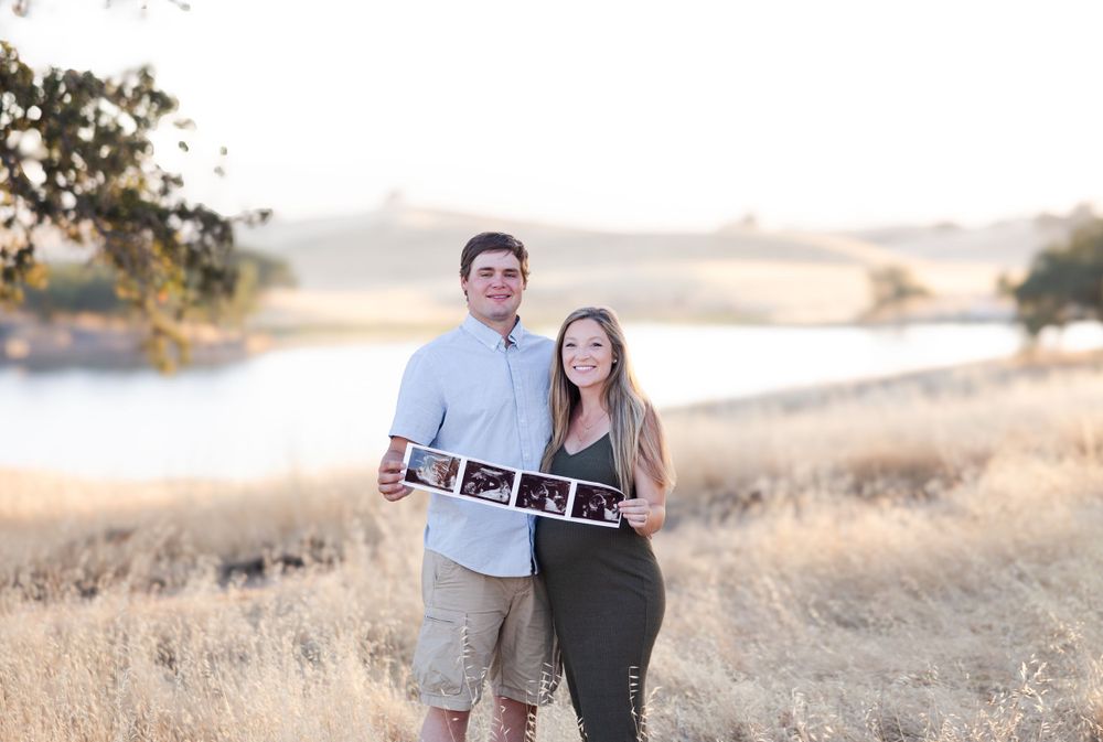Couple in casual attire holding ultrasound photos in golden field during sunset.