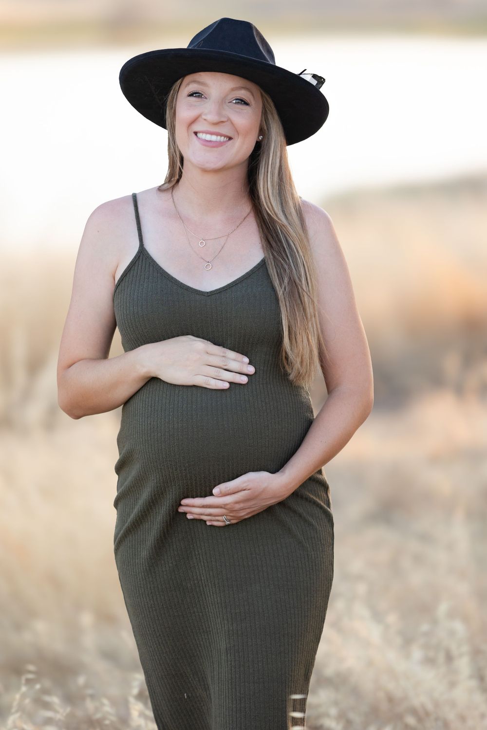 Expecting mother in olive green dress and black hat poses outdoors in a golden field at sunset.
