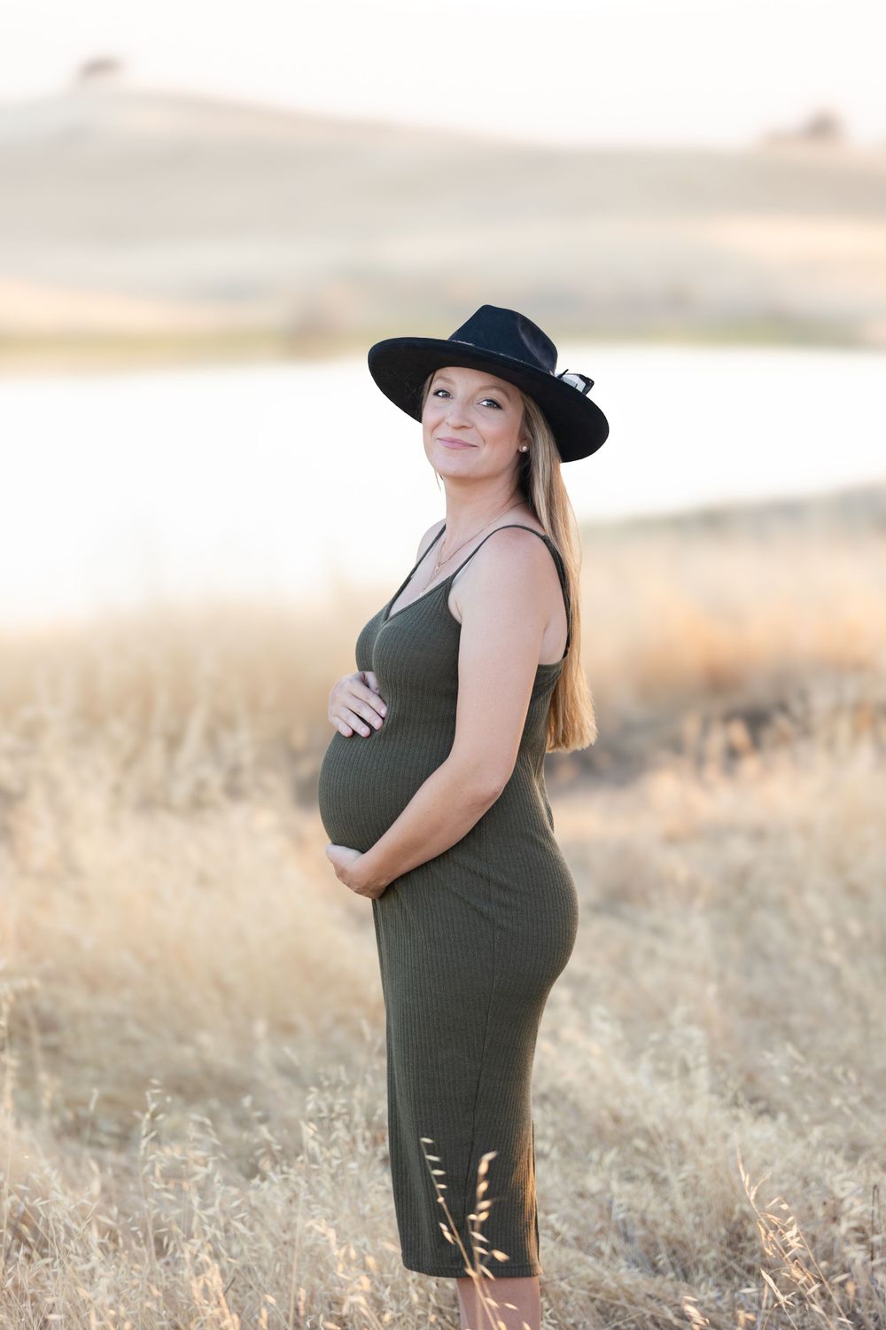 Expectant mother in black dress and hat poses in golden field during sunset maternity shoot.