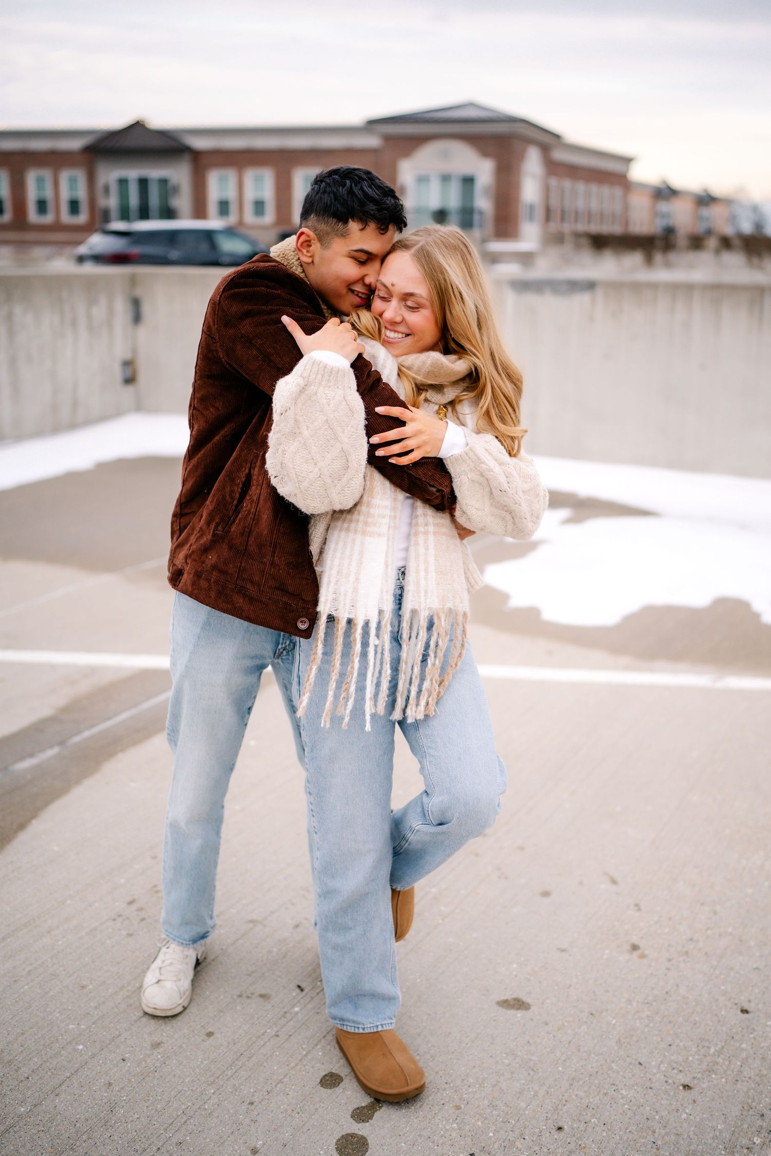 A couple in cozy winter sweaters embrace and laugh together on a snowy rooftop parking area.