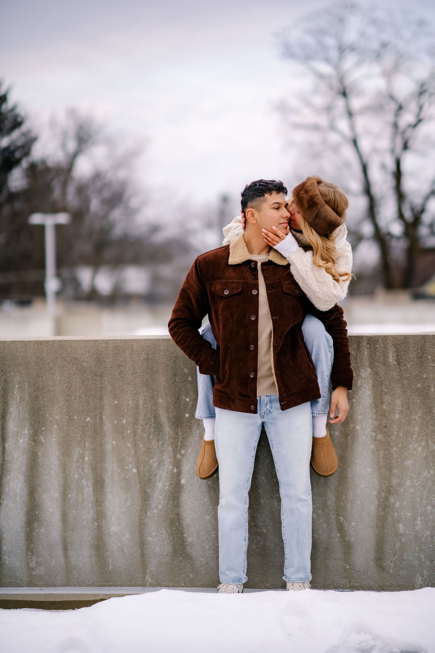 A romantic winter scene with a couple in casual wear standing against a concrete wall in the snow.