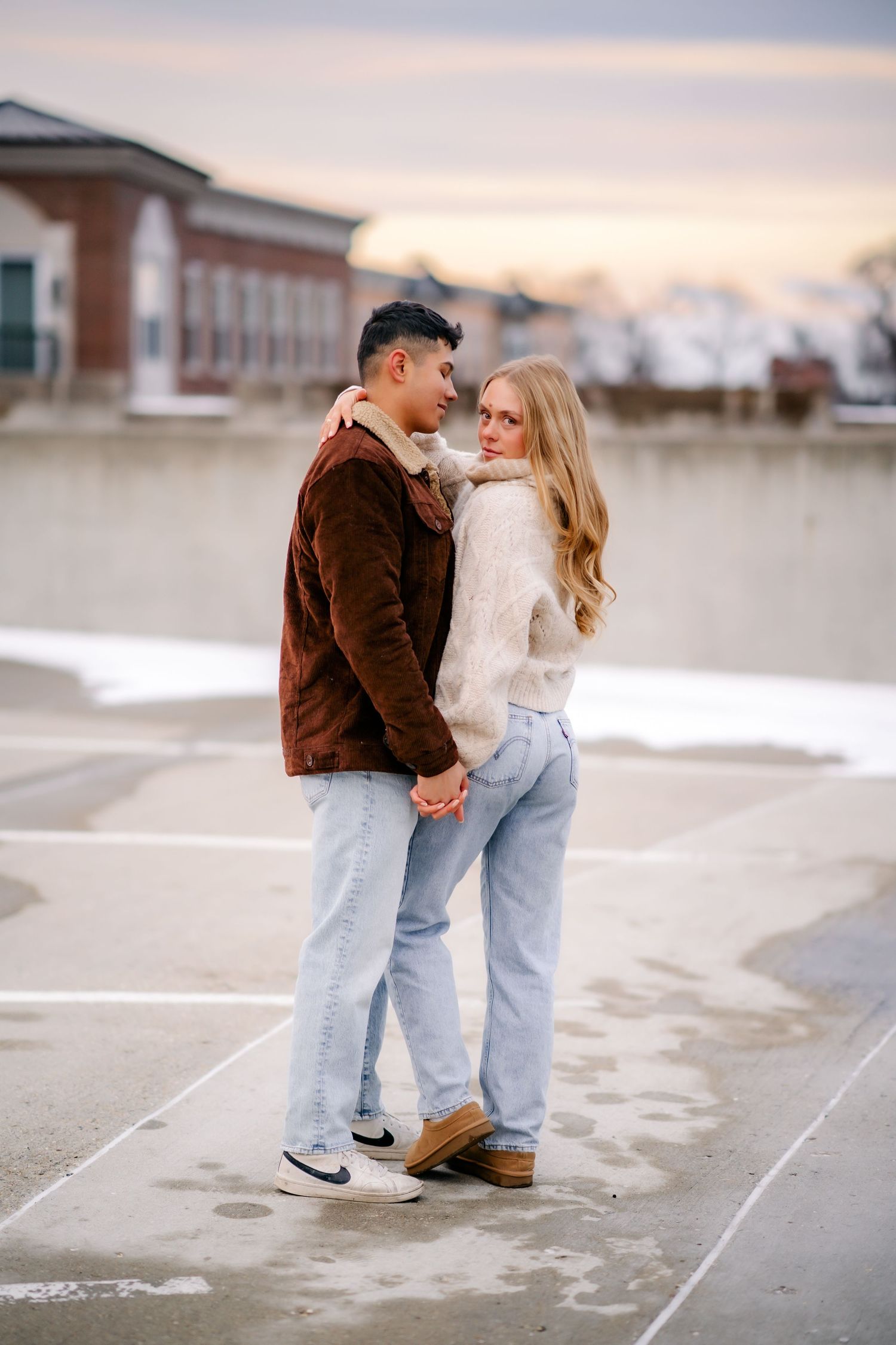 A couple in casual winter outfits holding hands and sharing a tender moment in a parking lot at sunset.