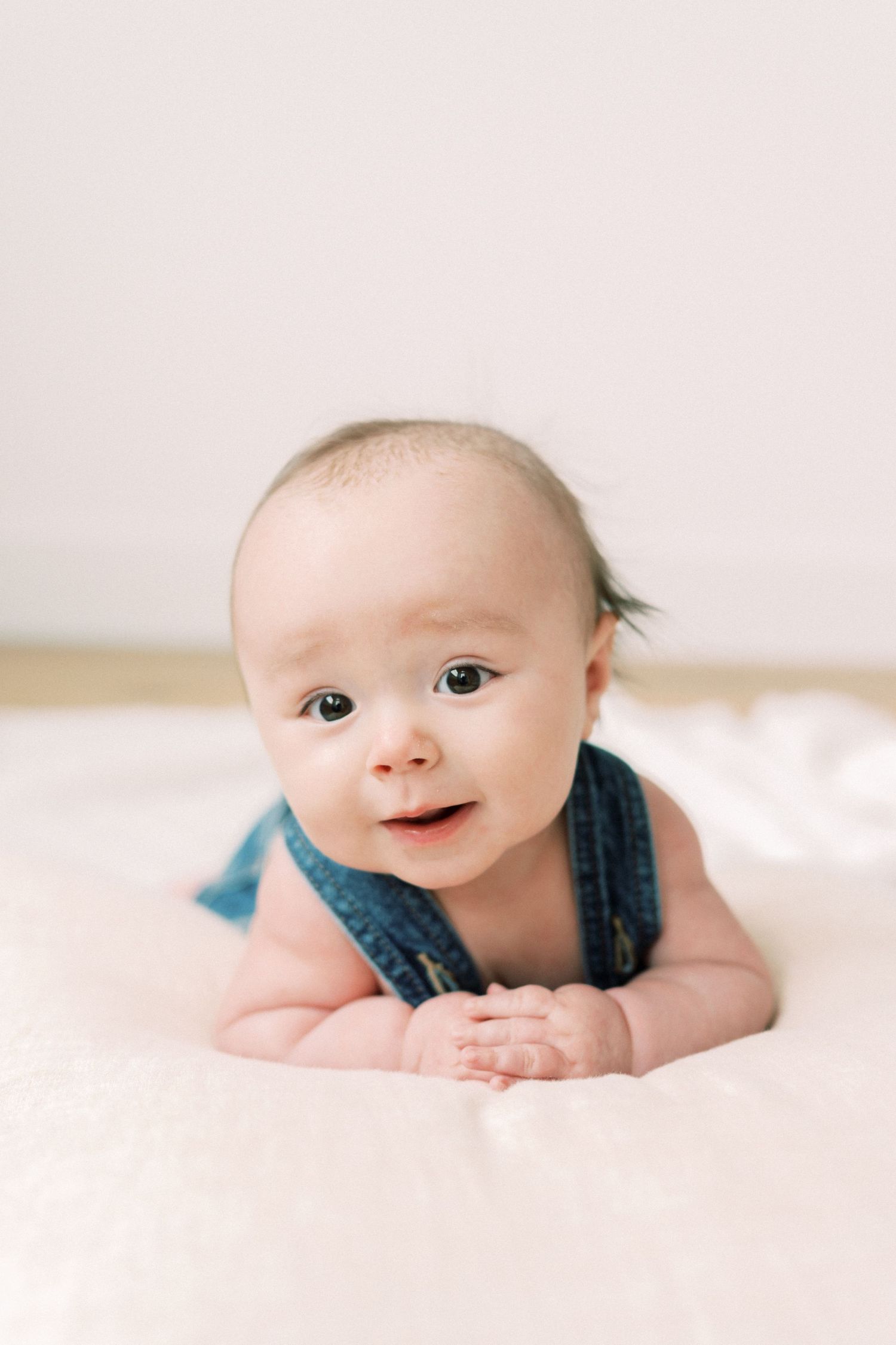 Adorable baby in denim overalls lying on white bedding with a sweet smile.