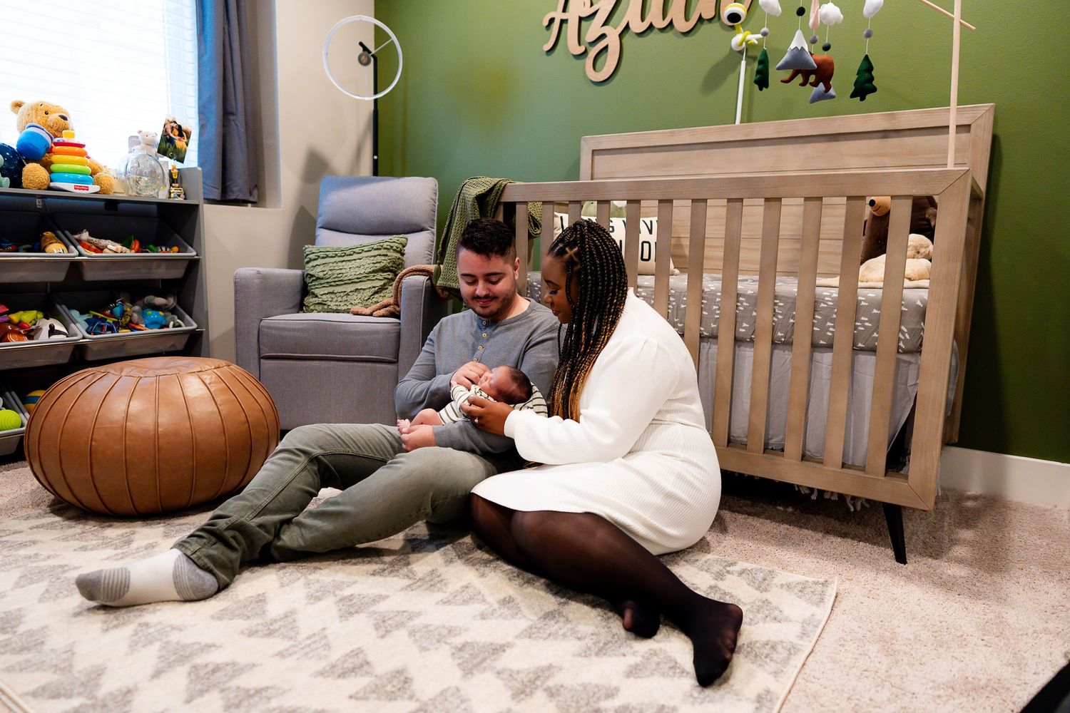 Parents lovingly hold a baby while sitting together on a plush gray rug in a modern nursery decorated with green walls during newborn photos