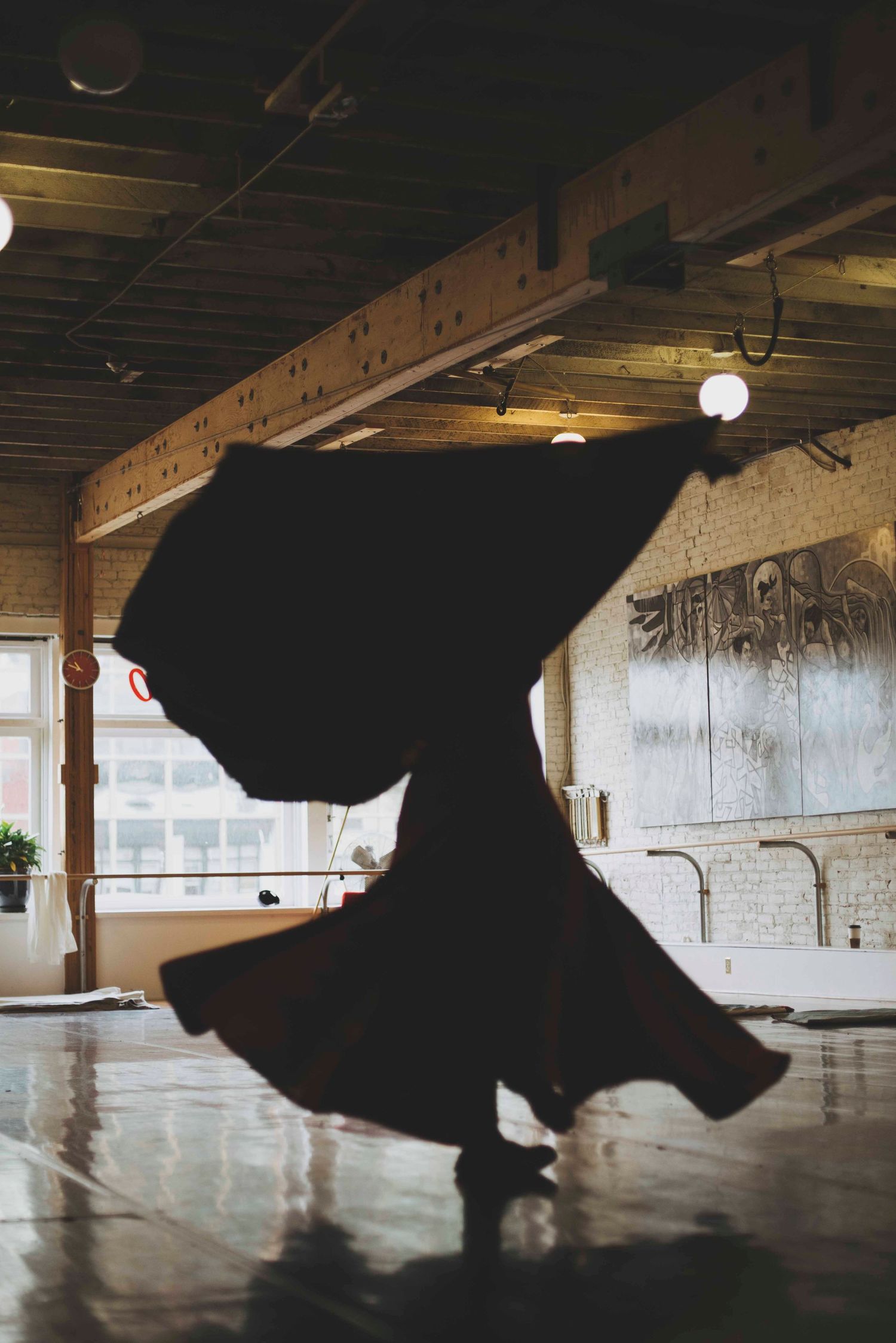 Dark silhouette of a dancer twirling in a flowy dress against industrial backdrop with exposed beams and globe lights.