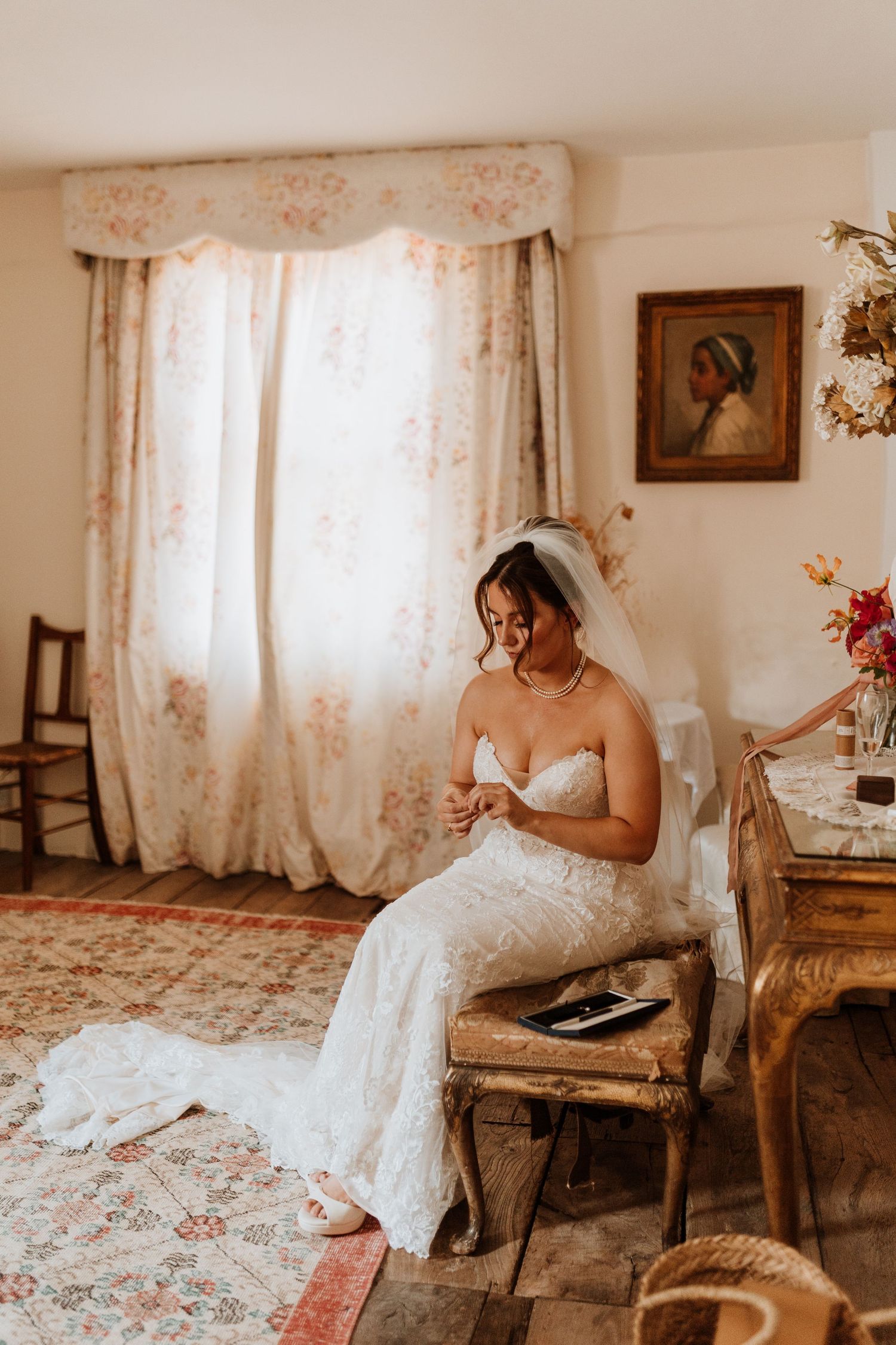 A bride in a white lace gown sits on a rustic chair by a window with floral curtains in a vintage-styled room at Lyde Court, Hereford.