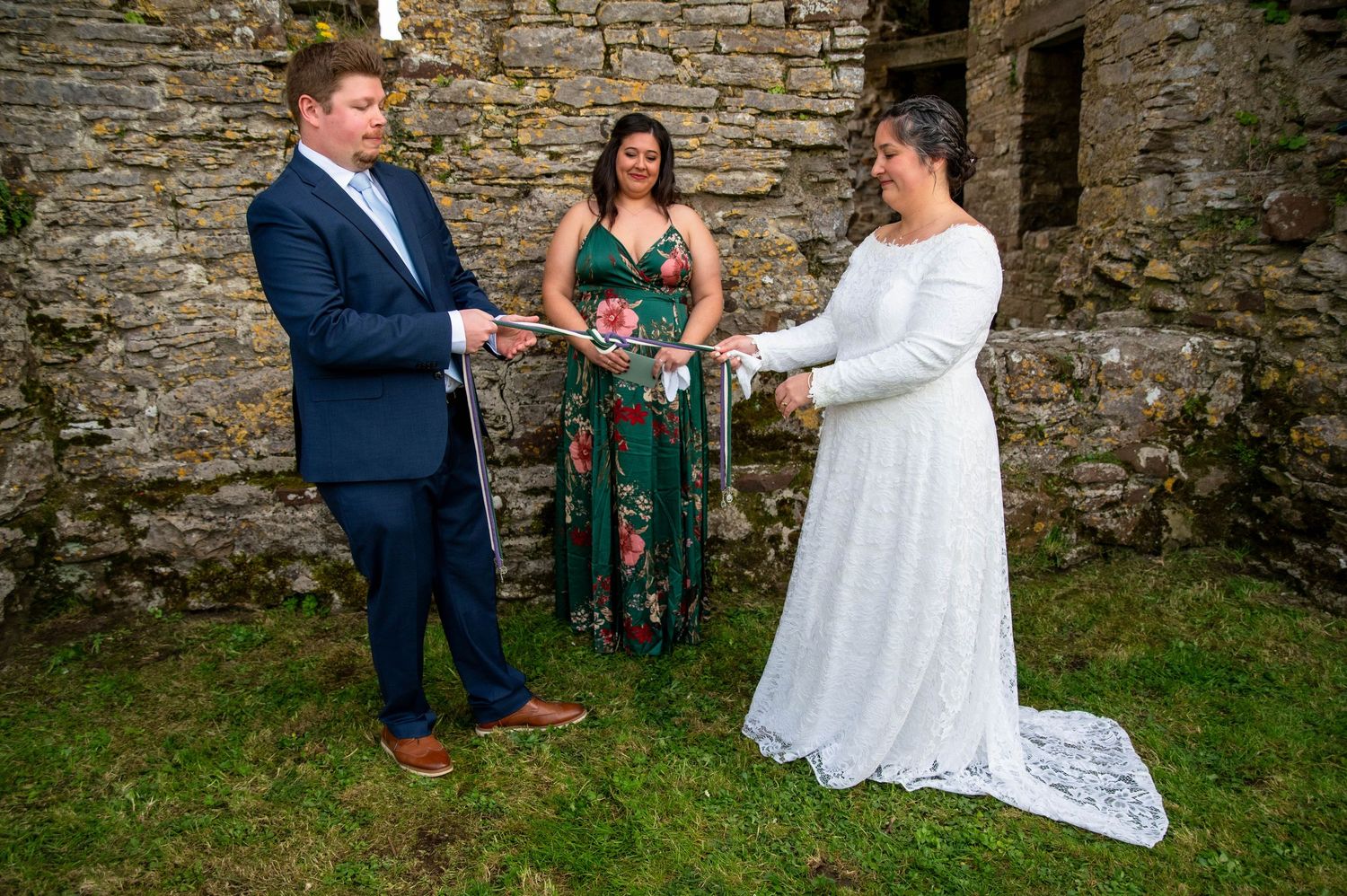 An outdoor wedding ceremony takes place beside a rustic stone wall with a person in green floral dress officiating.
