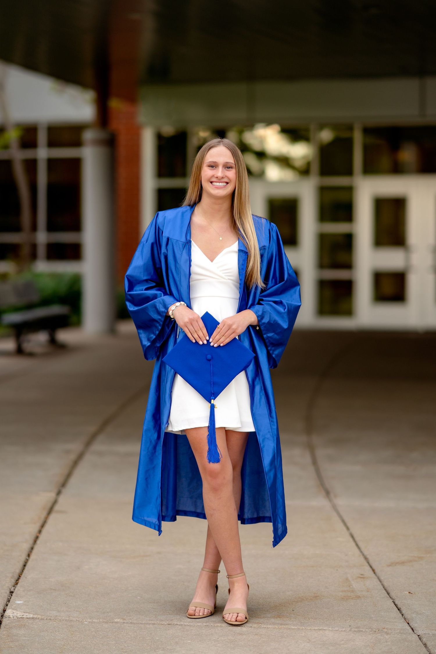 A graduates stands in front of a building wearing a bright blue graduation gown over a white dress.