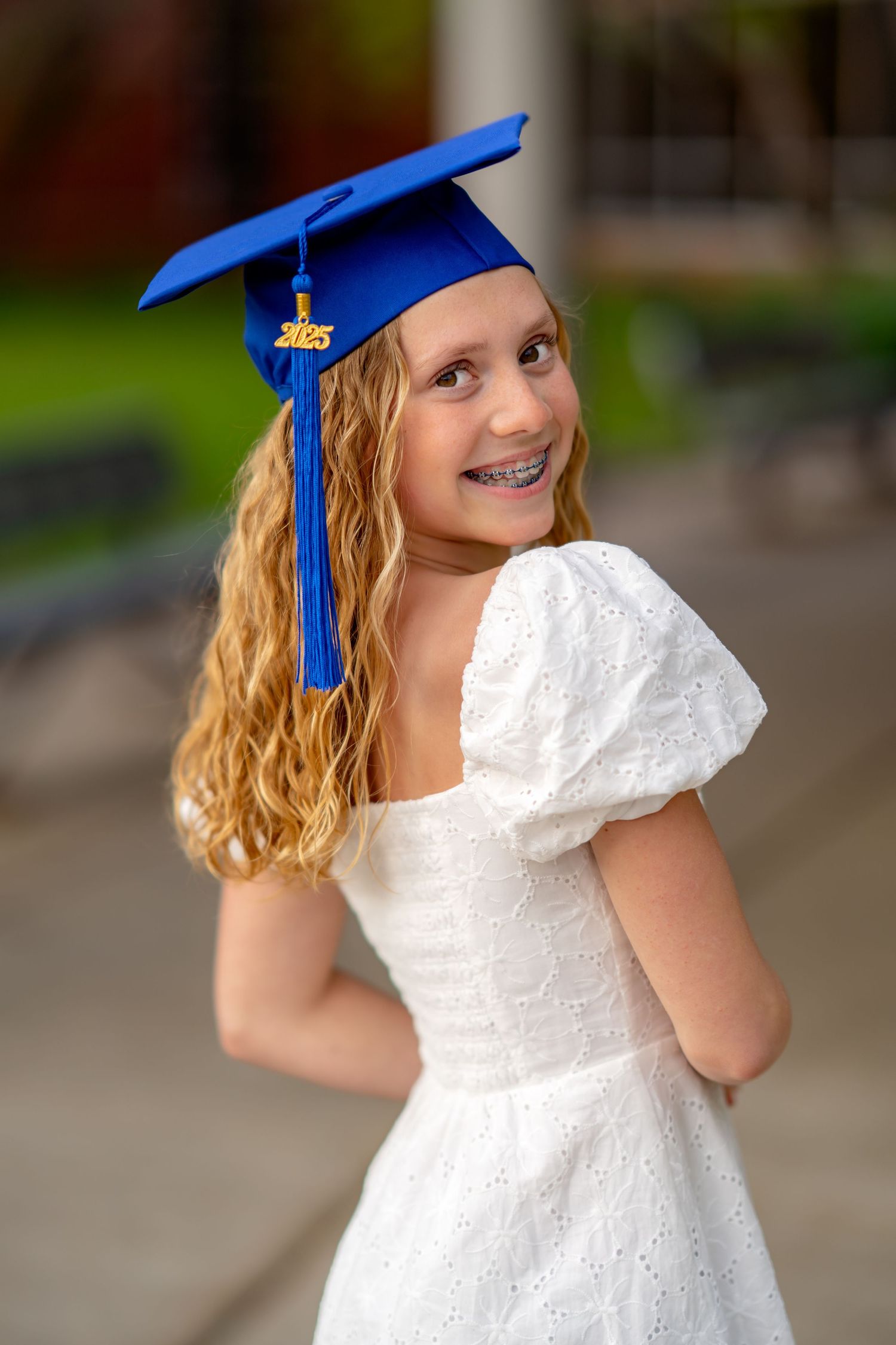 Graduate in white lace dress and bright blue cap and tassel smiles over shoulder in outdoor portrait.