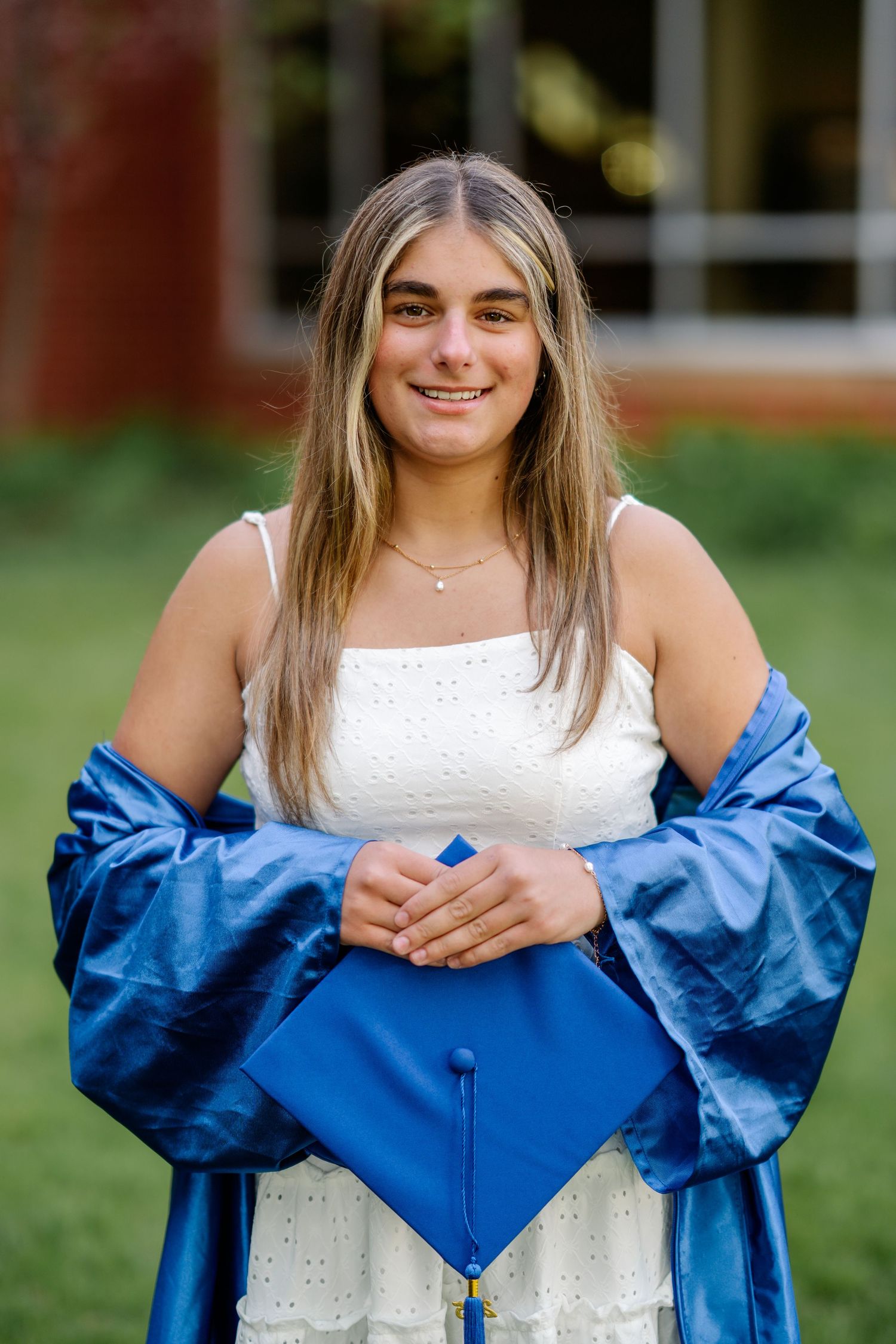 Graduate in white lace dress holds blue graduation gown and cap while posing outdoors on campus grounds.