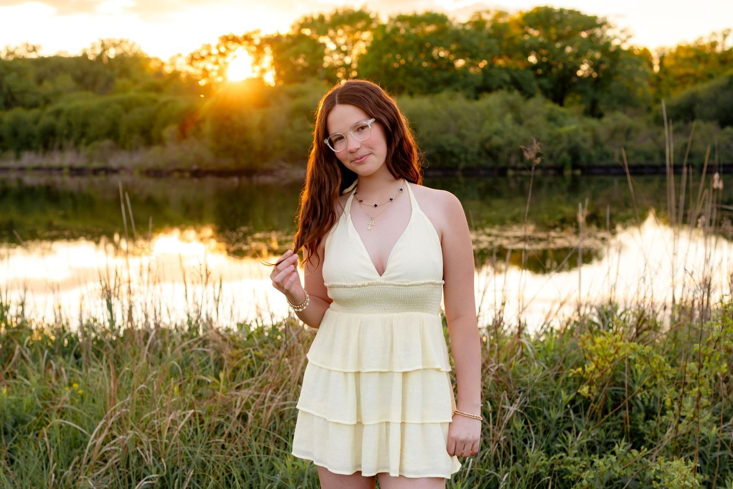 Person in white summer dress poses by scenic lake at sunset with golden light reflecting on water.
