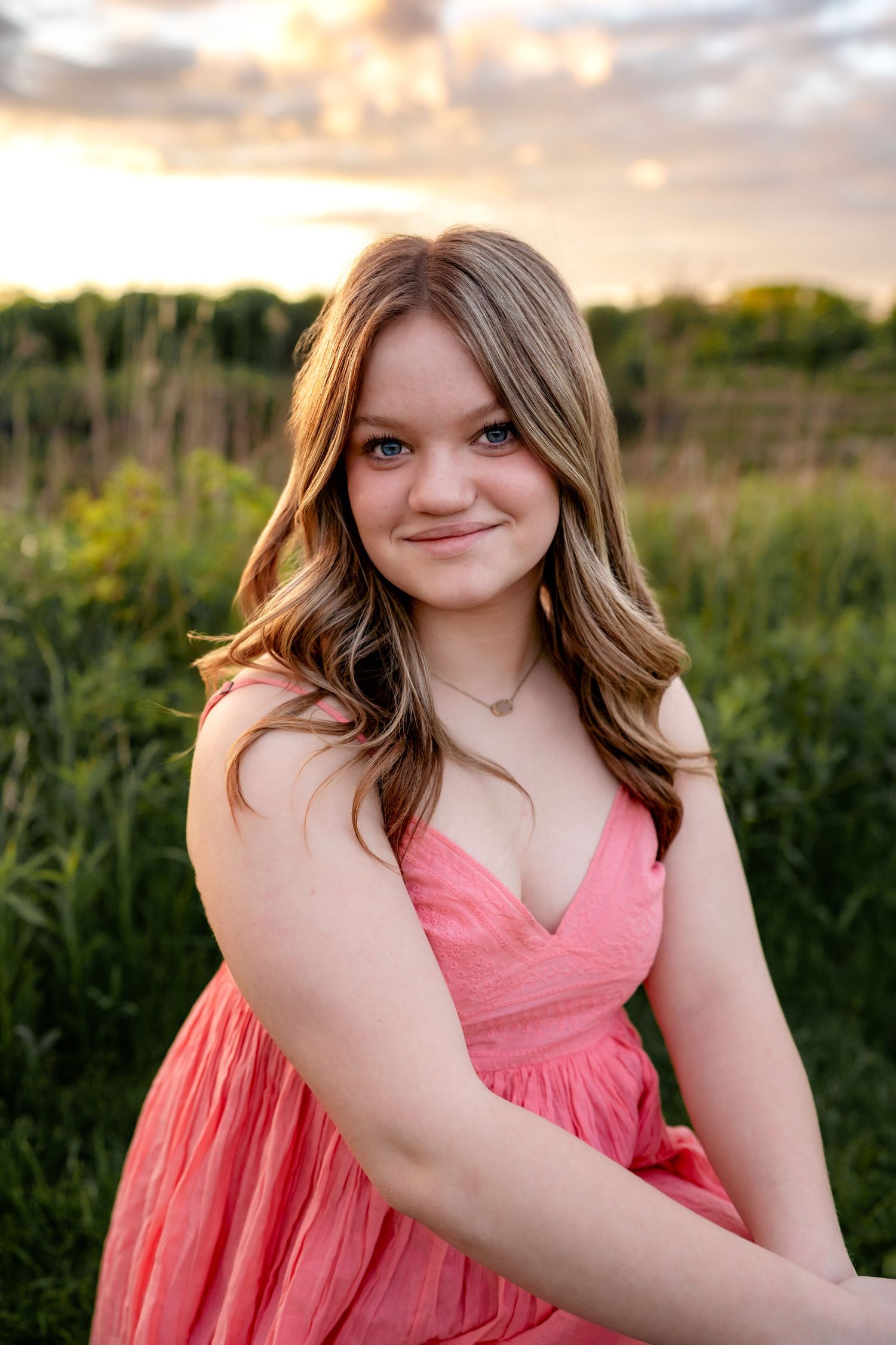 A person in a coral pink dress poses outdoors in a field at sunset with soft natural lighting.