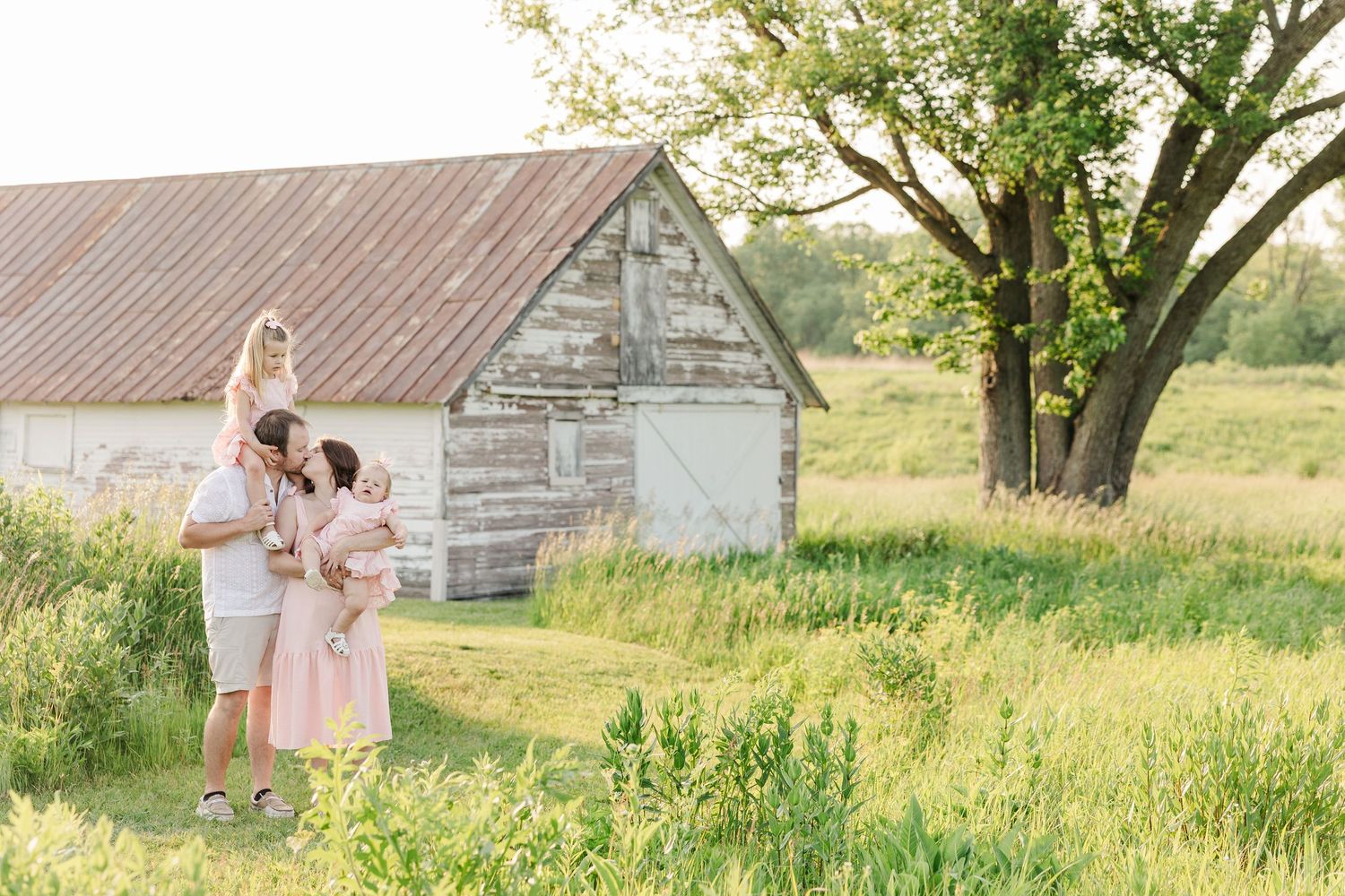 A family in pink and beige outfits stands together in a sunlit field beside a rustic weathered barn and large tree.