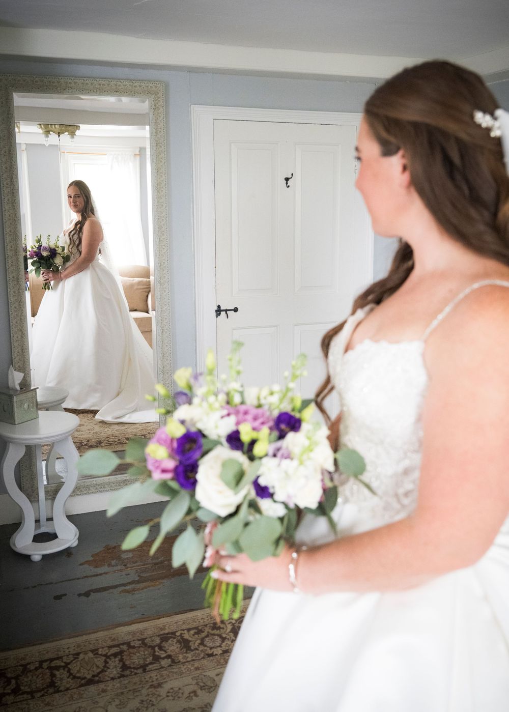 Wedding gown and flowers shown in bright natural light near white door and ornate mirror.