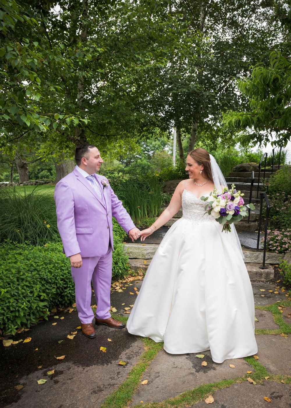 A couple shares a moment on their wedding day in a lush garden setting, with the bride in white and groom in lavender.