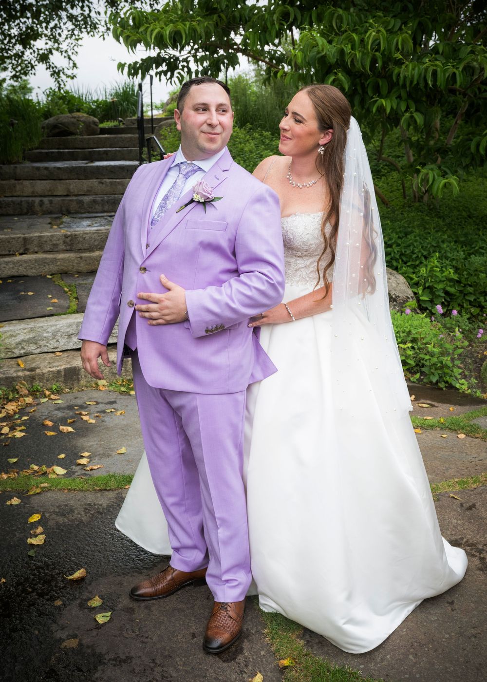 A newlywed couple poses outdoors on stone steps, with the groom wearing a lavender suit and bride in a white wedding gown.