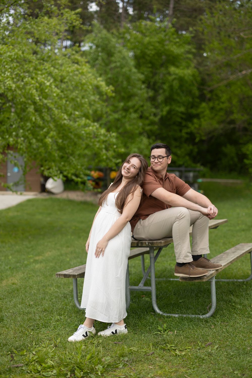 Couple in wedding attire share playful moments on park bench in lush green outdoor setting.