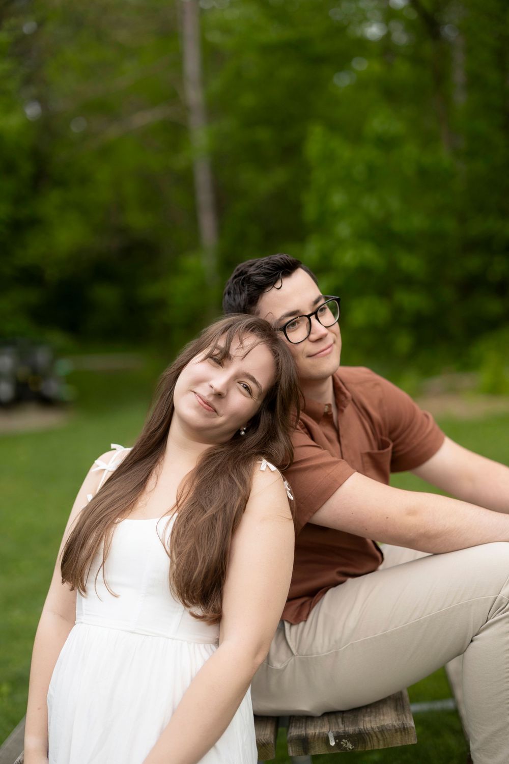 Couple relaxing outdoors in casual attire with long-haired person in white dress leaning on casual-dressed partner in glasses.
