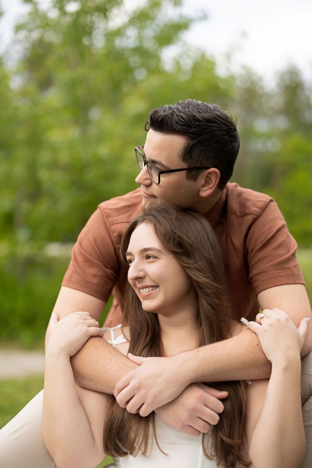 Couple sharing intimate moment outdoors with person in brown shirt embracing partner in white dress from behind.
