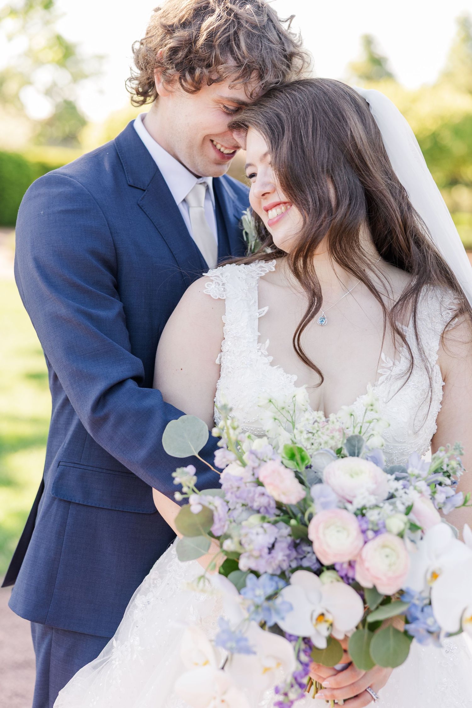 A happy wedding couple shares an intimate moment while holding a beautiful pastel bouquet of flowers in natural sunlight.